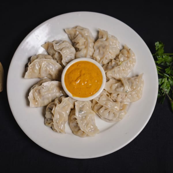 A white plate of steamed momos arranged around a small bowl of orange dipping sauce.