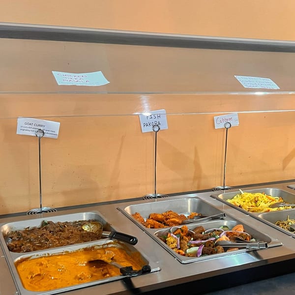 A buffet line with various Indian dishes including Goat Curry, Fish Pakora, and Cabbage.