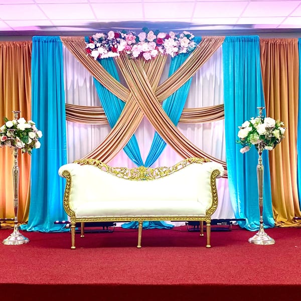 Elegant white and gold sofa on a red carpet stage, with blue and gold draped curtains and floral arrangements.
