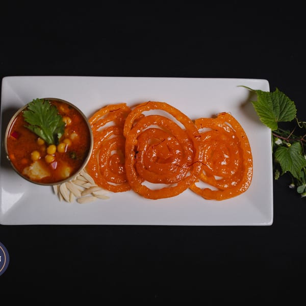 A white rectangular plate with three orange jalebis, a bowl of chickpea curry, and sliced almonds.
