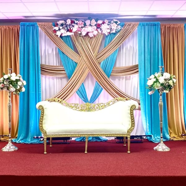 Elegant white and gold sofa on a red carpet stage, with blue and gold draped curtains and floral arrangements.