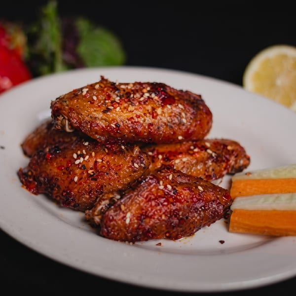 Spicy chicken wings with sesame seeds and chili flakes on a white plate with vegetable sticks.