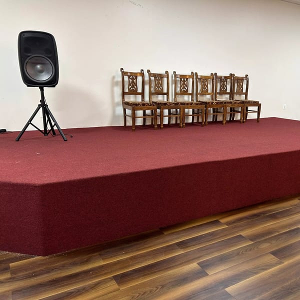 A black speaker on a stand and six wooden chairs with patterned seats on a red carpeted stage.
