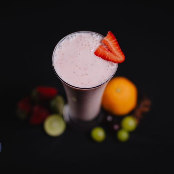 Strawberry milkshake in a tall glass, garnished with strawberry slices, surrounded by fruit on a dark background.