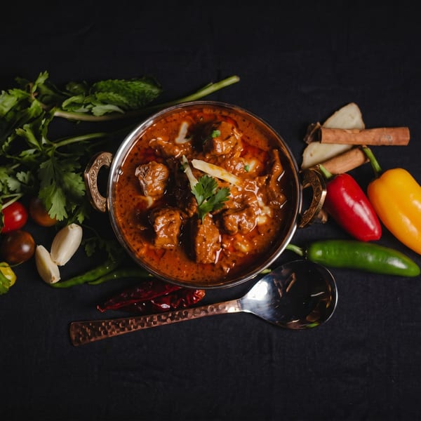 Indian curry in a metal bowl, surrounded by spices, herbs, and vegetables on a dark surface.