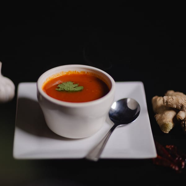Steaming red tomato soup with a cilantro garnish, spoon, garlic, and ginger on a white plate.