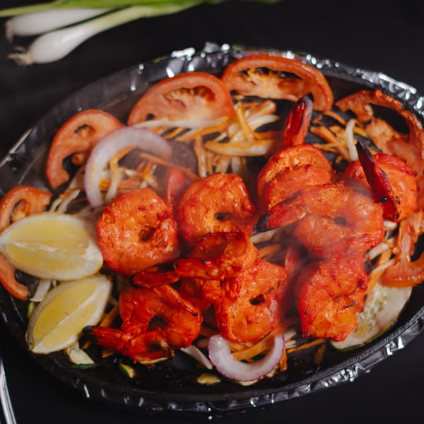Sizzling tandoori shrimp with sliced tomatoes, onions, and lemon wedges on a dark background.