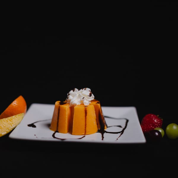 Orange flan with whipped cream and chocolate drizzle, garnished with fruit on a white plate.