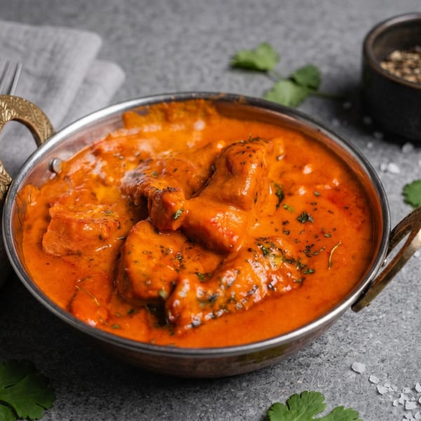 A metal bowl filled with vibrant orange butter chicken, garnished with herbs, on a gray surface.
