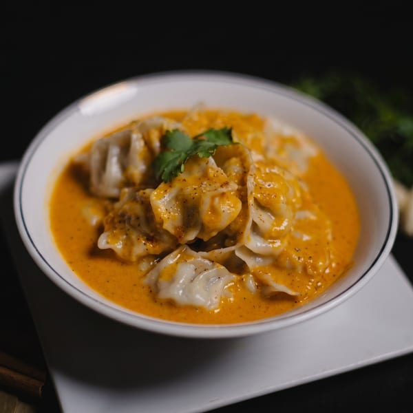 White bowl filled with momos in a rich orange sauce, garnished with fresh cilantro and pepper.
