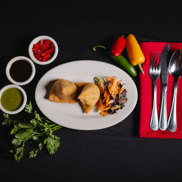 Two samosas with a side salad, three dipping sauces, and cutlery on a black background.