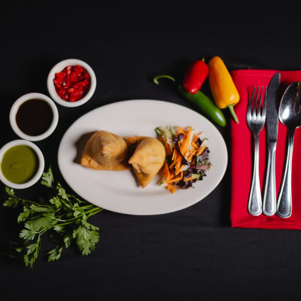 Two samosas with a side salad, three dipping sauces, and cutlery on a black background.