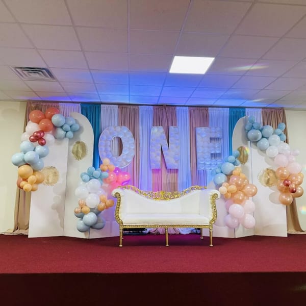 A stage with a white and gold ornate sofa, balloon garlands, and large "ONE" letters.
