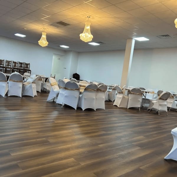 An empty banquet hall with white tables and chairs, gold sashes, and chandeliers.