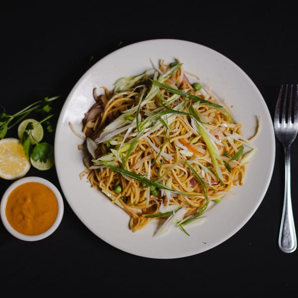 Noodles with green onions, peas, and other vegetables, served with a side of sauce and lemon.