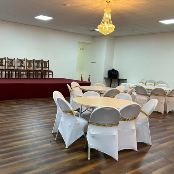 Empty banquet hall with round tables, white chair covers, a red stage, and a gold chandelier.