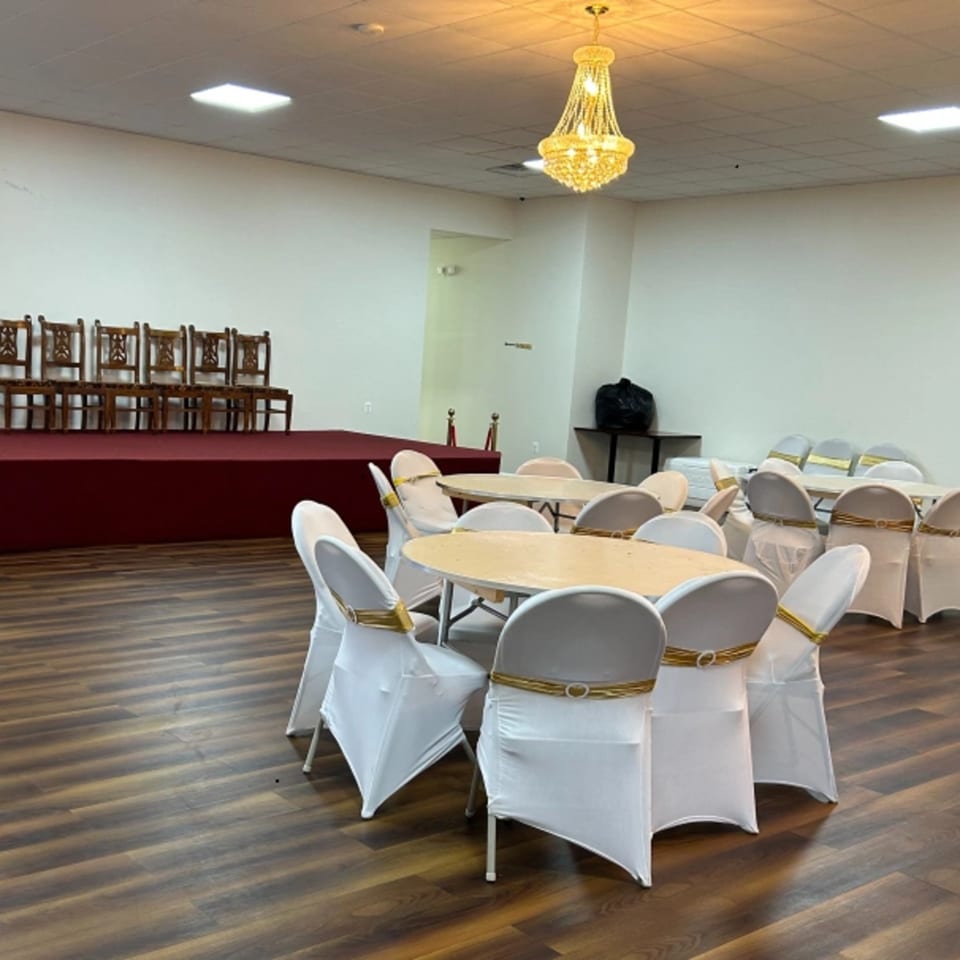 Empty banquet hall with round tables, white chair covers, a red stage, and a gold chandelier.
