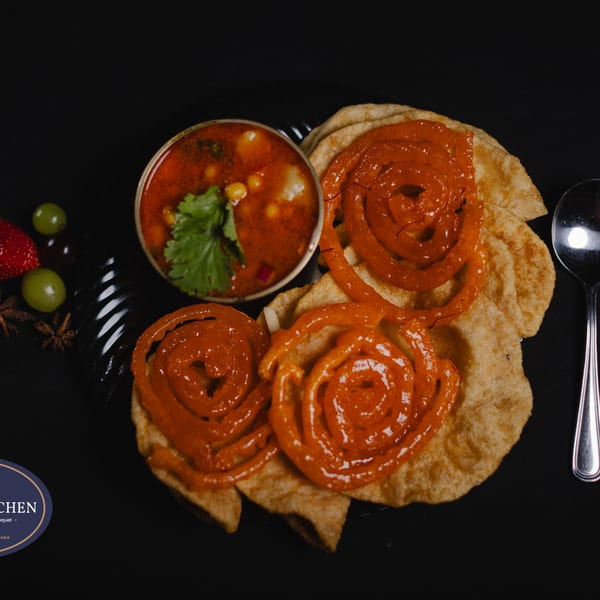 Chana masala with cilantro, jalebi, and puri bread on a black plate with fruit and spices.
