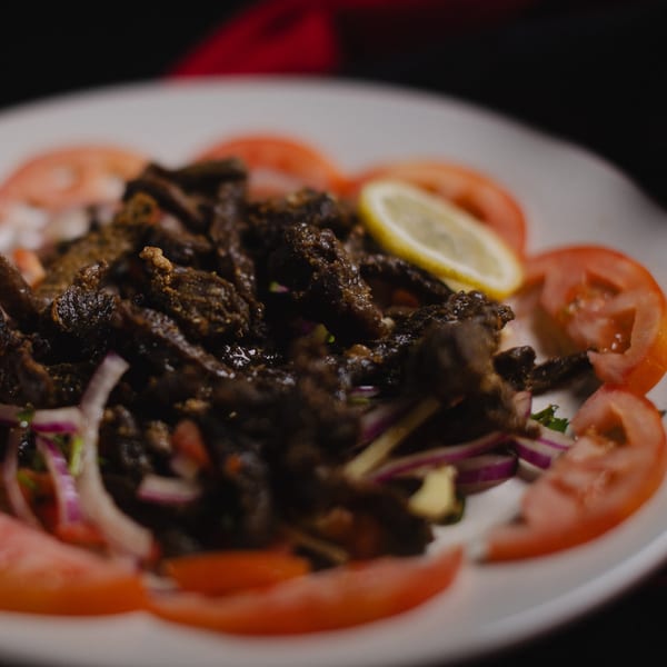 Close-up of dark, seasoned meat strips on a white plate, garnished with tomato slices and red onion.