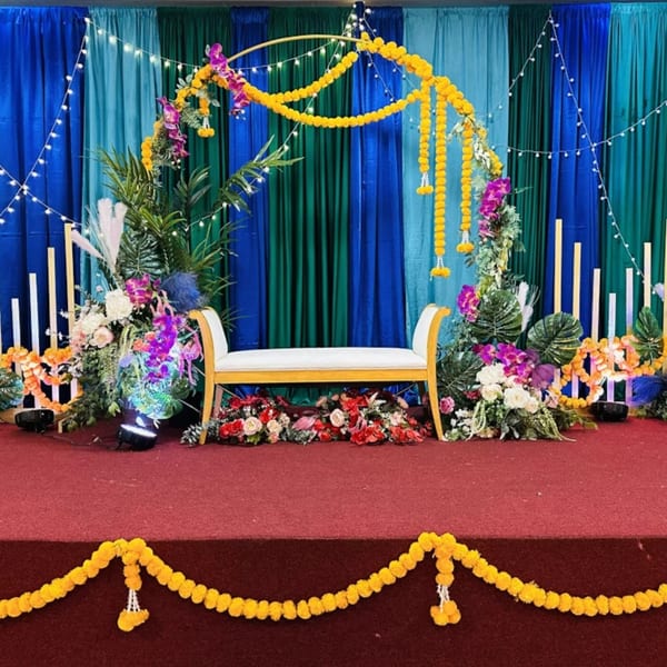 A decorated stage with a white bench, floral arrangements, and marigold garlands against blue and green drapes.