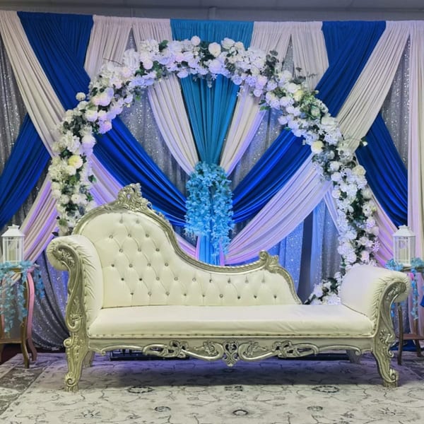 Ornate white and silver chaise lounge in front of a blue, white, and silver draped backdrop with floral arch.