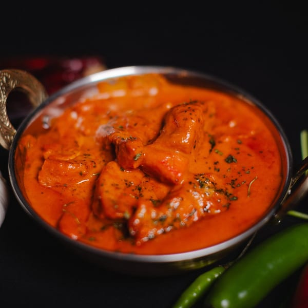 Close-up of a metal bowl filled with vibrant orange butter chicken, garnished with herbs.