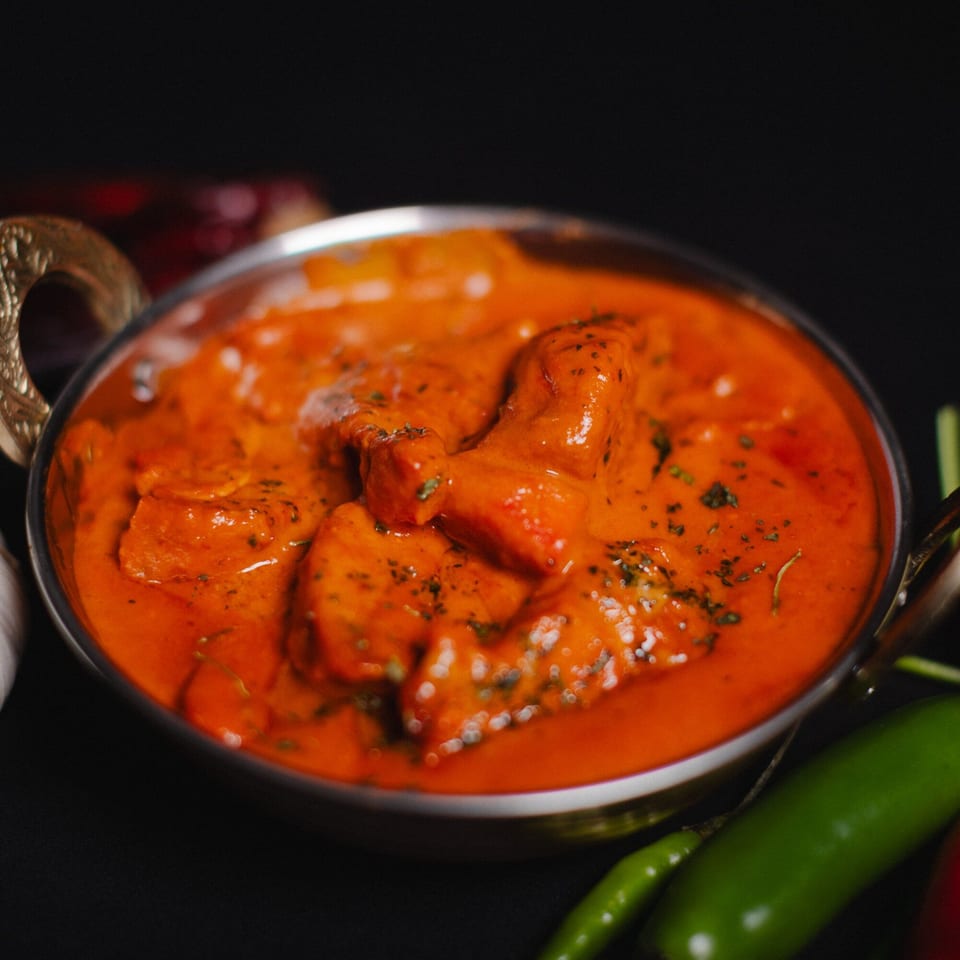Close-up of a metal bowl filled with vibrant orange butter chicken, garnished with herbs.