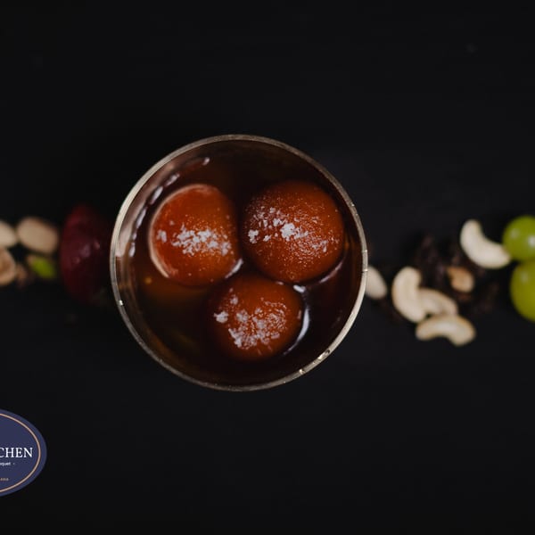 Three gulab jamuns in a metal cup, garnished with coconut flakes, surrounded by nuts and fruit.