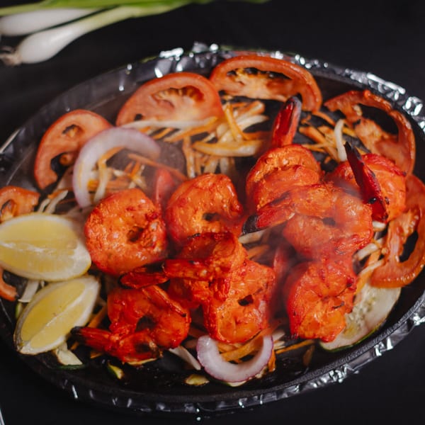 Sizzling red shrimp with sliced tomatoes, onions, and lemon wedges on a dark platter.