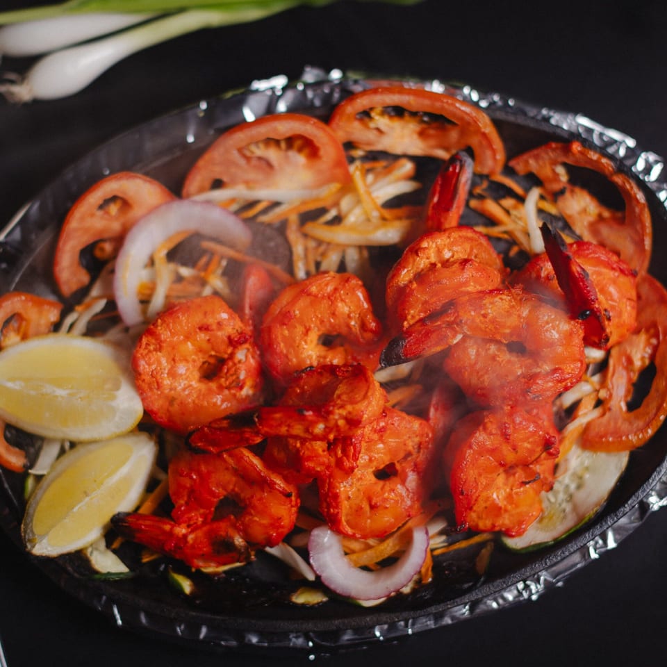 Sizzling red shrimp with sliced tomatoes, onions, and lemon wedges on a dark platter.