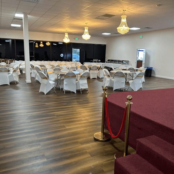 A large banquet hall with round tables, white chairs with gold sashes, and a red carpeted stage.