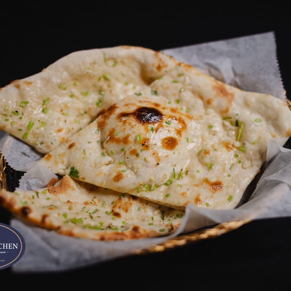 Close-up of golden-brown naan bread, garnished with green herbs, served in a basket with parchment paper.